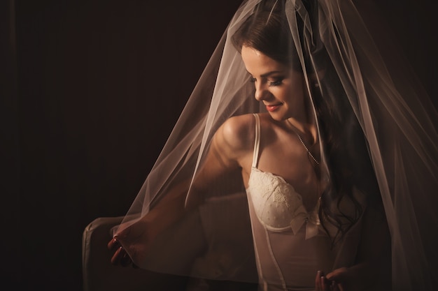 Foto preparativi mattutini. chiuda sul colpo della sposa elegante in lingerie bianca in posa sotto il primo piano di velo su sfondo scuro.