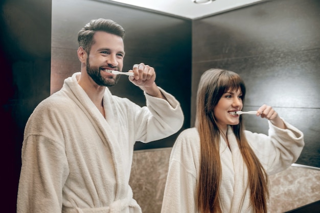 Morning procedures. Young couple in bath robes brushing teeth together