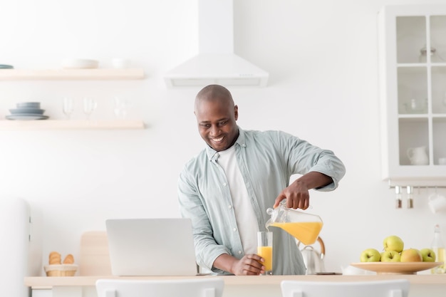 Morning routine. Happy mature black man having video chat with family on laptop computer, pouring juice on the kitchen table and looking at pc screen, empty space