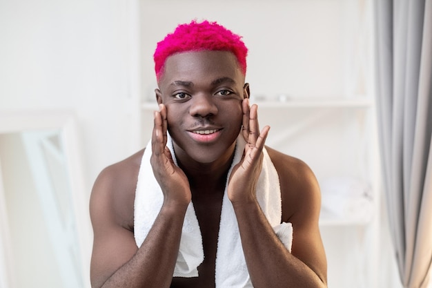 Morning routine Metrosexual black man Facial care Hygiene procedure Handsome shirtless african guy with bright pink hair towel on shoulder touching face looking at camera bathroom interior