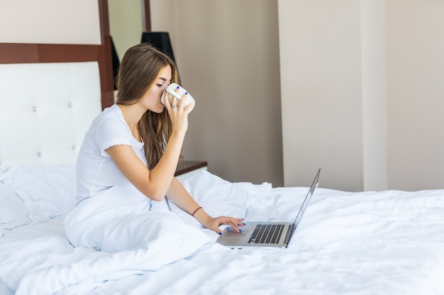 La routine del mattino. bella giovane donna che beve caffè e naviga in internet sul suo laptop mentre è seduta a letto e sorride alla telecamera