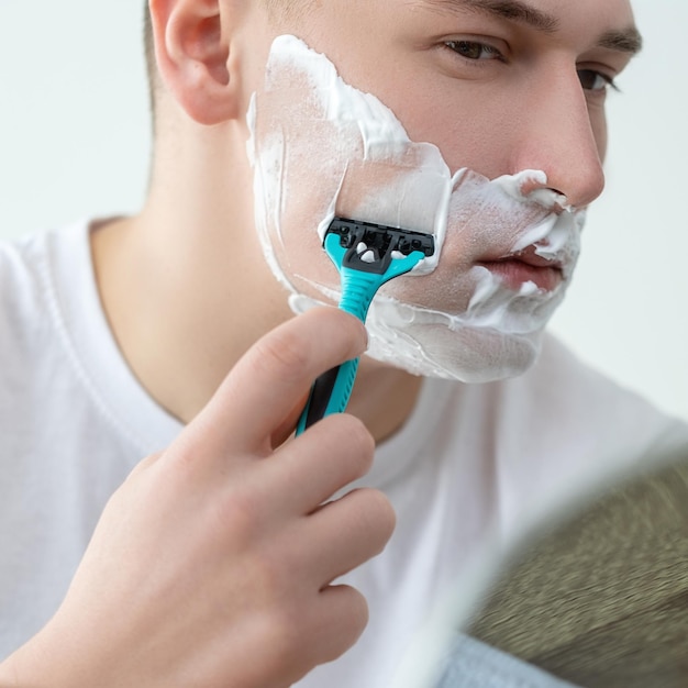 Morning shave Selfcare routine Facial treatment Concentrated serious attractive young man with shaving foam on face looking in mirror using razor on white background