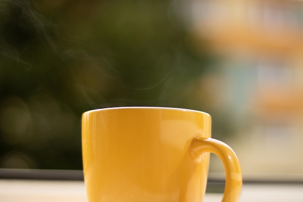 Morning tea on the windowsill from which steam comes out. Yellow cup with a drink near