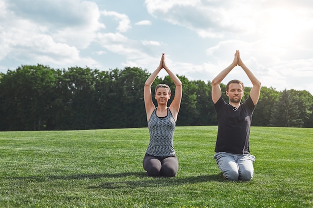 Morning time woman and man meditating together outdoors while sitting on a green lawn in