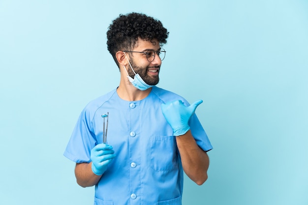 Moroccan dentist man holding tools isolated on blue background pointing to the side to present a product