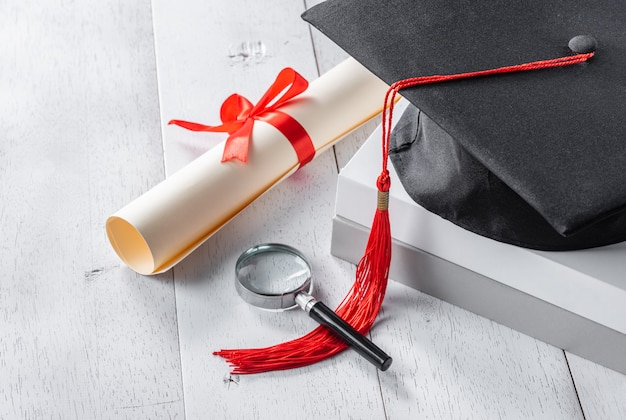 Photo mortarboard, magnifying glass and diploma tied with red ribbon on white wooden table