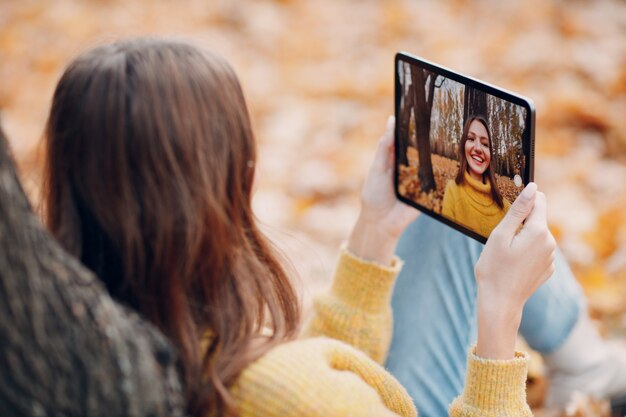 MOSCOW, RUSSIA - OCTOBER 19, 2021: Young woman model take selfie photo with tablet pc in autumn park with yellow foliage maple leaves. Fall season fashion