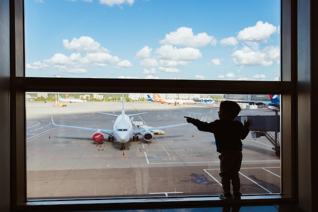 Moskou Rusland 07 14 22 Kleine jongen draagtas wachten op het instappen naar de vlucht in de transithal van de luchthaven en kijken door het raam naar het vertrek van het vliegtuig Actieve gezinslevensstijl reizen door de lucht met kind