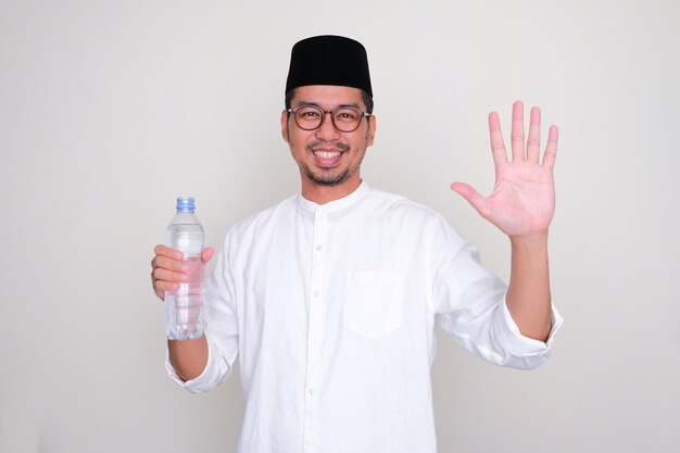 Moslem Asian man smiling and showing five fingers while holding a drinking water bottle