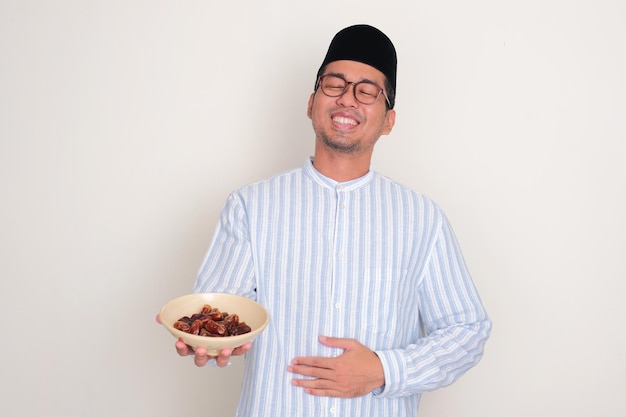Moslim Aziatische man die zijn buik met een opgeluchte uitdrukking aanraakt terwijl hij dadelfruit vasthoudt