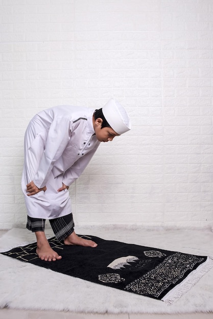 Moslim kind in wit overhemd, sarong en kalotje doet rakooh, buigend op gebedsmat