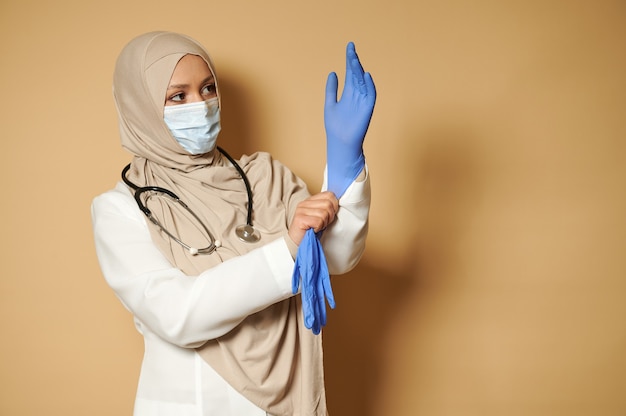 Moslim vrouwelijke arts met bedekt hoofd het dragen van medische beschermende handschoenen poseren voor voorzijde op beige oppervlak met kopie ruimte