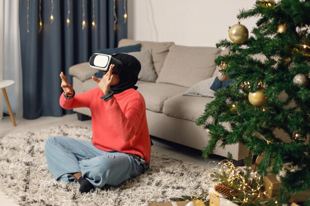 Moslimmeisje in zwarte hijab zit in de buurt van de kerstboom en draagt een virtual reality-bril