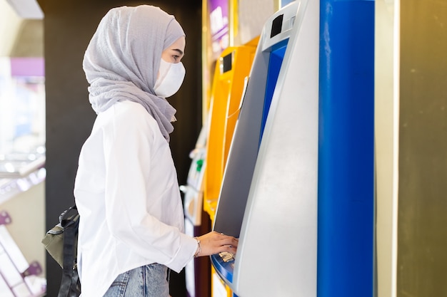 Moslimvrouw die medisch masker draagt om besmettingsvirus te voorkomen dat ATM-machine gebruikt om geld op stadsstraat op te nemen.