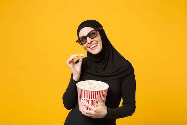 Moslimvrouw in hijab zwarte kleding 3d imax bril kijken film film houd emmer popcorn, eet popcorn geïsoleerd op gele muur portret. mensen levensstijl concept. .