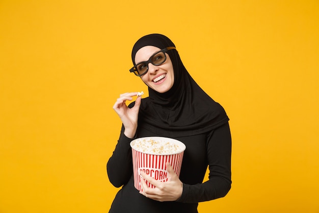 Moslimvrouw in hijab zwarte kleding 3d imax bril kijken film film houd emmer popcorn, eet popcorn geïsoleerd op gele muur portret. Mensen levensstijl concept. .