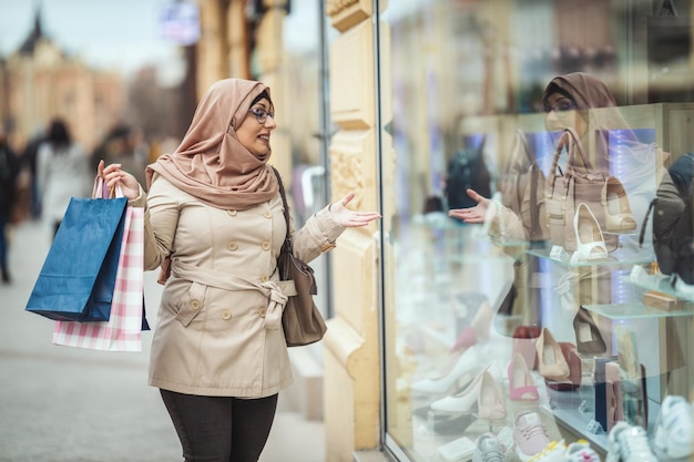 Moslimvrouw van middelbare leeftijd die hijab met een blij gezicht draagt, loopt in een stedelijke omgeving en gaat naar de winkel met papieren gekleurde tassen.