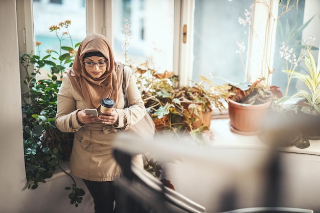 Moslimvrouw van middelbare leeftijd die hijab met een blij gezicht draagt, staat bij het raam en tipt berichten op haar smartphone.