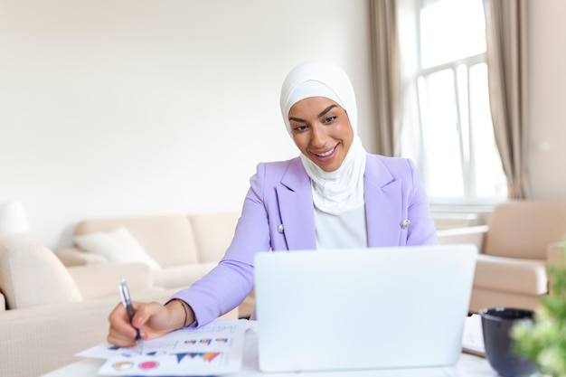 Moslimvrouw werkt in thuiskantoor Arabische vrouw gekleed in religieuze sluier werkt op haar laptop, kijkt naar het scherm en schrijft notities