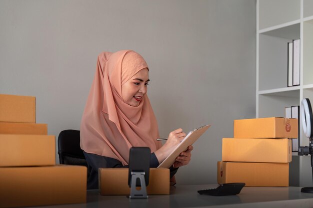 Moslimvrouwen die thuis online verkopen met een doos Online verkopen met een doos om bestelling van een klant te accepteren KMO bedrijfsidee Parcelbezorging Moslimvrouw die thuis werkt met smartphone en laptop.