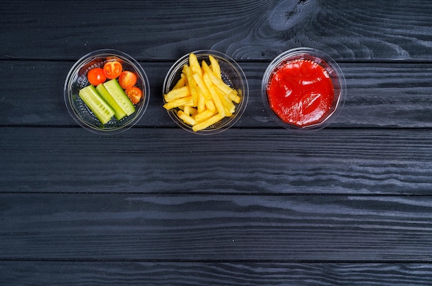 Most popular fast food meal French fries with ketchup and vegetables on dark wooden background top view copy space