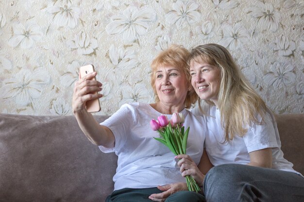 Mother and adult blonde daughter in white t-shirts are smiling and making selfie with bouquet of pink tulips at home. Mother's day concept