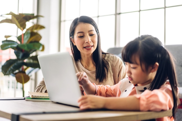 Мать и азиатский ребенок маленькая девочка учится и смотрит на портативный компьютер, делая домашнее задание, изучая знания с помощью системы электронного обучения онлайн. Детская видеоконференция с учителем-наставником дома