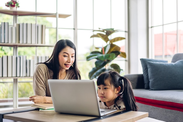 Mother and asian kid little girl learning and looking at laptop computer making homework studying knowledge with online education e-learning system.children video conference with teacher tutor at home