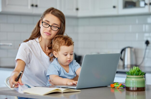 Mother baby and book with laptop for remote work at kitchen for reading ideas or editing in family home Woman infant and computer for creativity in notes freelance or thinking for media company