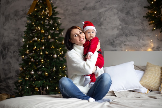 Mother and baby in santa red hat smile with Christmas trees in the interior of the house