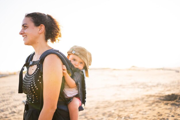 Mother carrying a baby on her back in a baby carrier outdoors during sunset