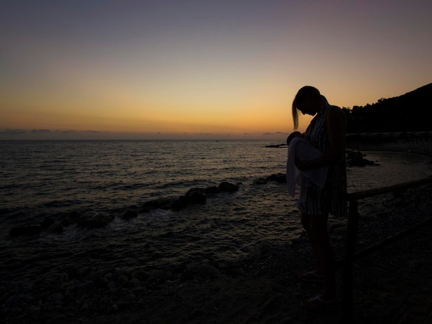 Mother carrying daughter at beach against sky during sunset