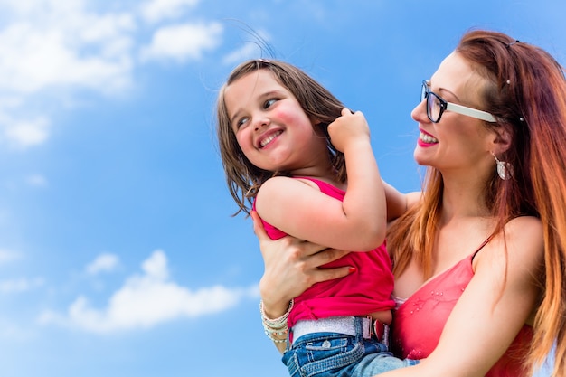 Madre che porta la figlia sul braccio sotto un cielo estivo azzurro