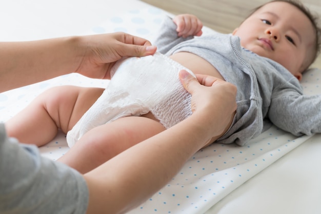 Mother changing diaper for her son on white bed in bedroom