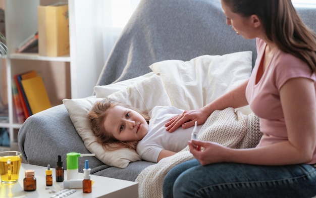 Mother checks daughter's temperature on thermometer. There medicines on table.