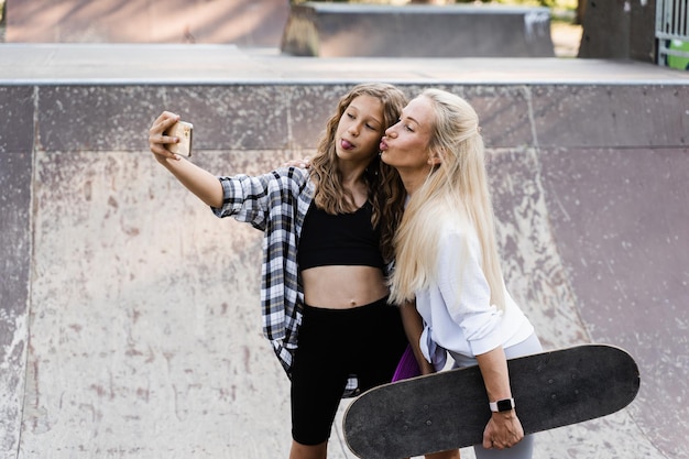 Mother and child daughter with skate and penny board is making selfie on phone in skate park Extreme lifestyle Family of mom and her kid spend time together