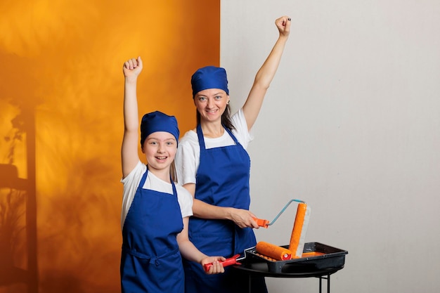 Mother and child having fun painting walls, using orange paint\
color to change apartment room. feeling happy about house\
renovation with roller paintbrush and decorating tools at\
home.