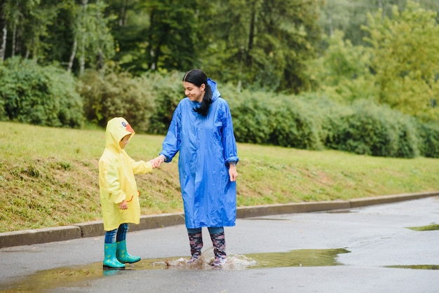 母と子が雨の中で遊んで、ブーツとレインコートを着ている