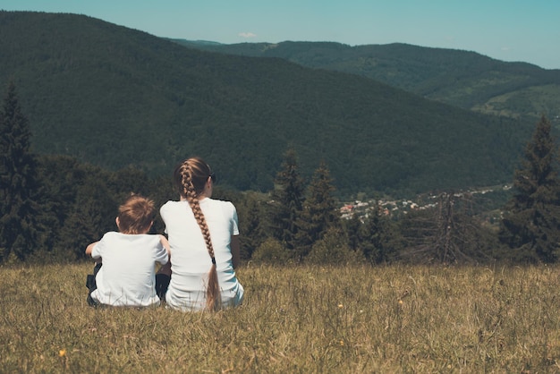 Mother and child sitting in a meadow and look at nature Sister and younger brother People mountains in the background Back view