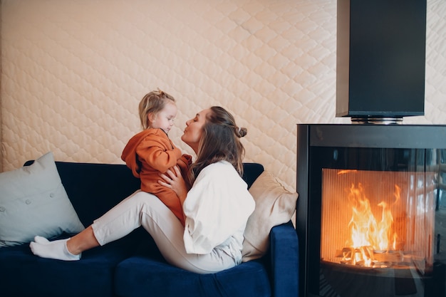 Mother and child sitting and playing on sofa near fireplace. Mom and baby. Parent and little kid relaxing at home. Family having fun together.