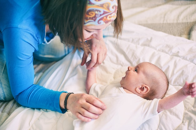 Mother and child on a white bed. Mom and baby boy in diaper playing in sunny bedroom. Mom makes gymnastics for her newborn baby.