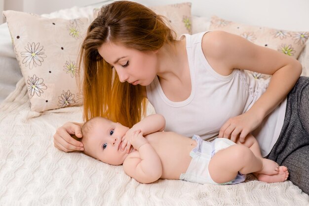 Mother and child on a white bed mom and baby girl in diaper playing in sunny bedroom