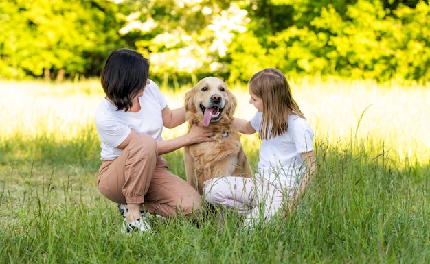 Mother and cute daughter hugging with adorable golden retriever dog outdoors