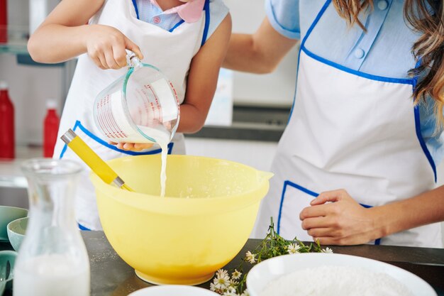 Mother and daughter in apron pouring warm milk in bowl when making cookie dough