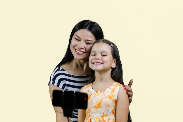 어머니와 딸이 파스텔 노란색에 고립된 막대기를 사용하여 스마트폰으로 셀카를 만들고 있습니다.