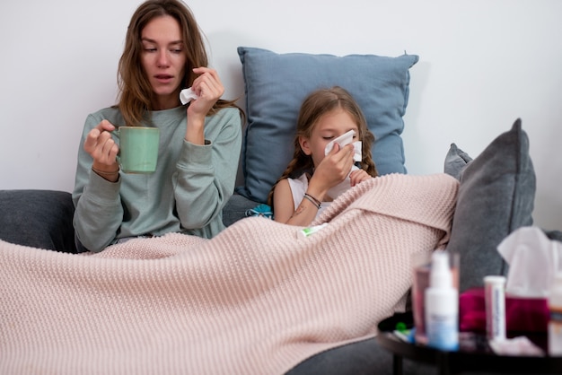 Mother and daughter are sick together sitting on the couch under the plaid.