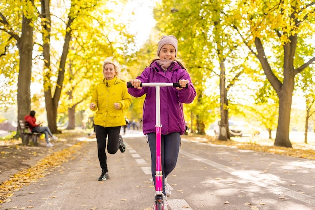 Mother and daughter are walking in the autumn in the park. Little girl is riding a scooter