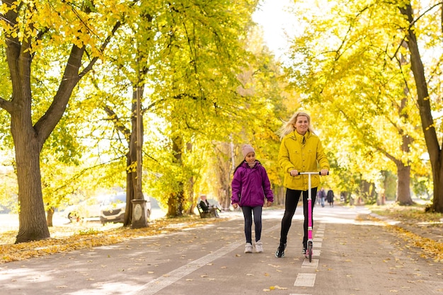 엄마와 딸이 공원에서 가을을 걷고 있습니다. 여자가 스쿠터를 타고 있다