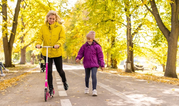 Photo mother and daughter are walking in the autumn in the park. woman is riding a scooter