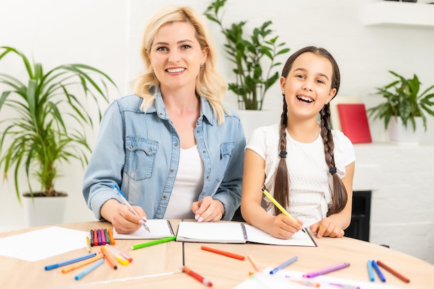 Mother and daughter beautiful and happy make a card for mother's day sitting together at the table with paper and pencils.